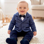 Elegancki garniturek do chrztu dla niemowlaka składa się z marynarki w niebieską pepitkę, spodni granatowych, białego koszulo-body, muszki w kolorze granatowym oraz kaszkietu również w kolorze granatowym.