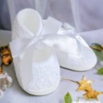 Białe koronkowe balerinki dla niemowlaka do chrztu z dużą atłasową kokardą.