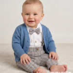 Eleganckie ubranko do chrztu dla chłopca: spodnie w beżową kratkę, biała koszula z muszką i niebieski sweterek rozpinany.