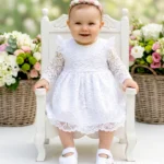 Biała sukienka do chrztu z koronki z długim rękawem i ozdobnymi patkami na ramionach.